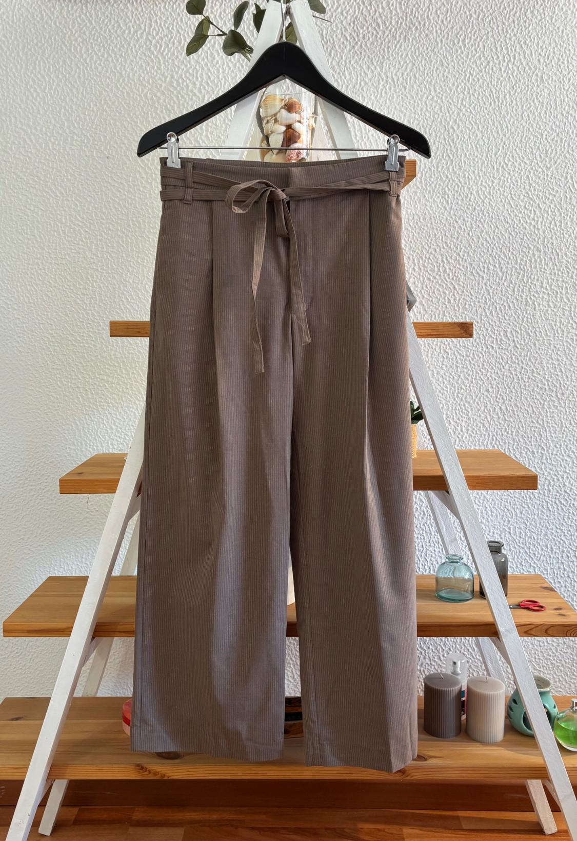 Light brown fabric trousers with a belt, hanging on a black hanger over a decorative wooden ladder.