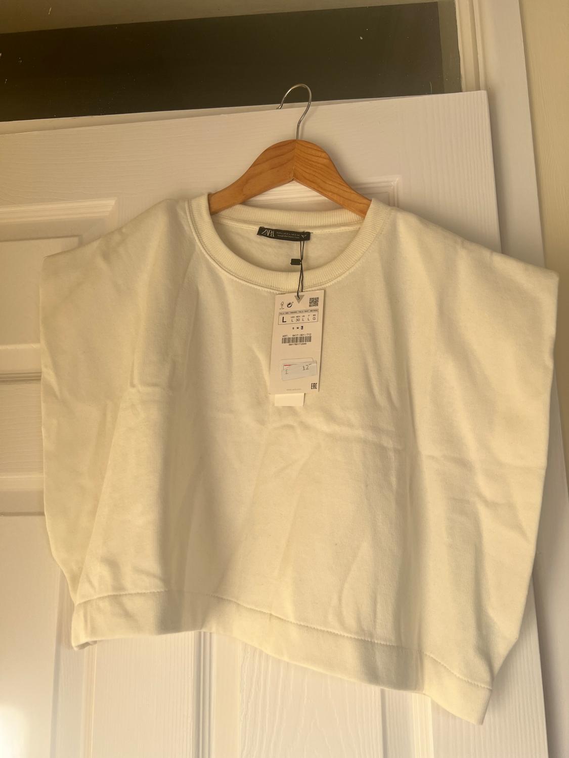 White cropped t-shirt hanging on a wooden hanger over a white door. Visible tag with product details.