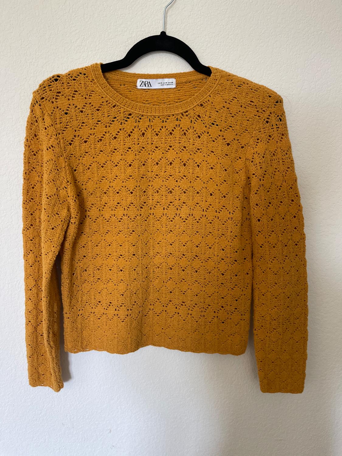 Mustard-coloured open knit jumper hanging on a black hanger. Visible label with text: "ZARA". Plain white background.