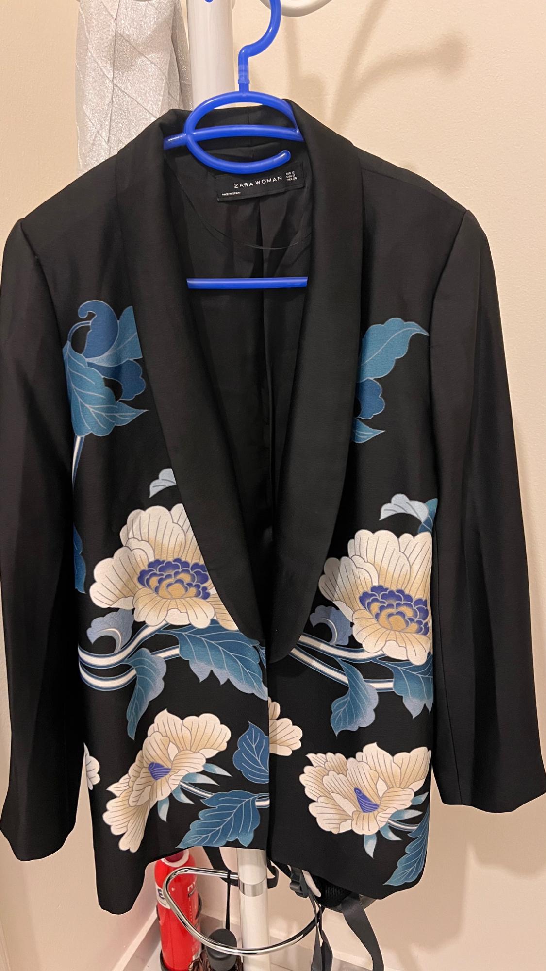 Black jacket with a blue and beige floral print, hanging on a blue hanger. Visible label: "ZARA WOMAN". Neutral background.
