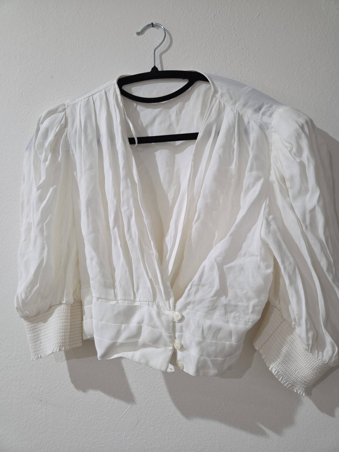 White short-sleeve blouse with a V-neck, button-front closure, and gathered cuffs, hanging on a black hanger against a white wall.