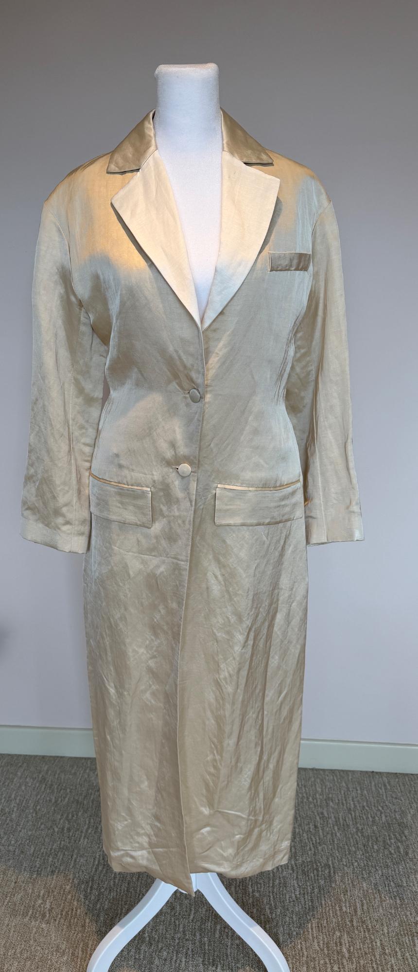 Long beige coat on a white mannequin, with wide lapels and two front buttons. Light gray background and carpeted floor.