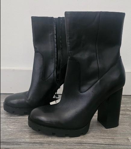 Black ankle boots with chunky heels and treaded soles, with a side zip, on a wooden floor in front of a white wall.