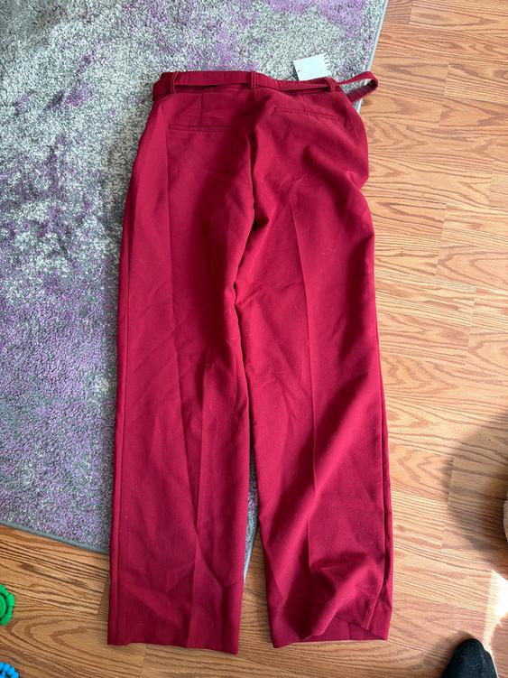 Red fabric pants on a wooden floor and purple carpet. Visible tag at the waist. Natural light illuminates the scene.