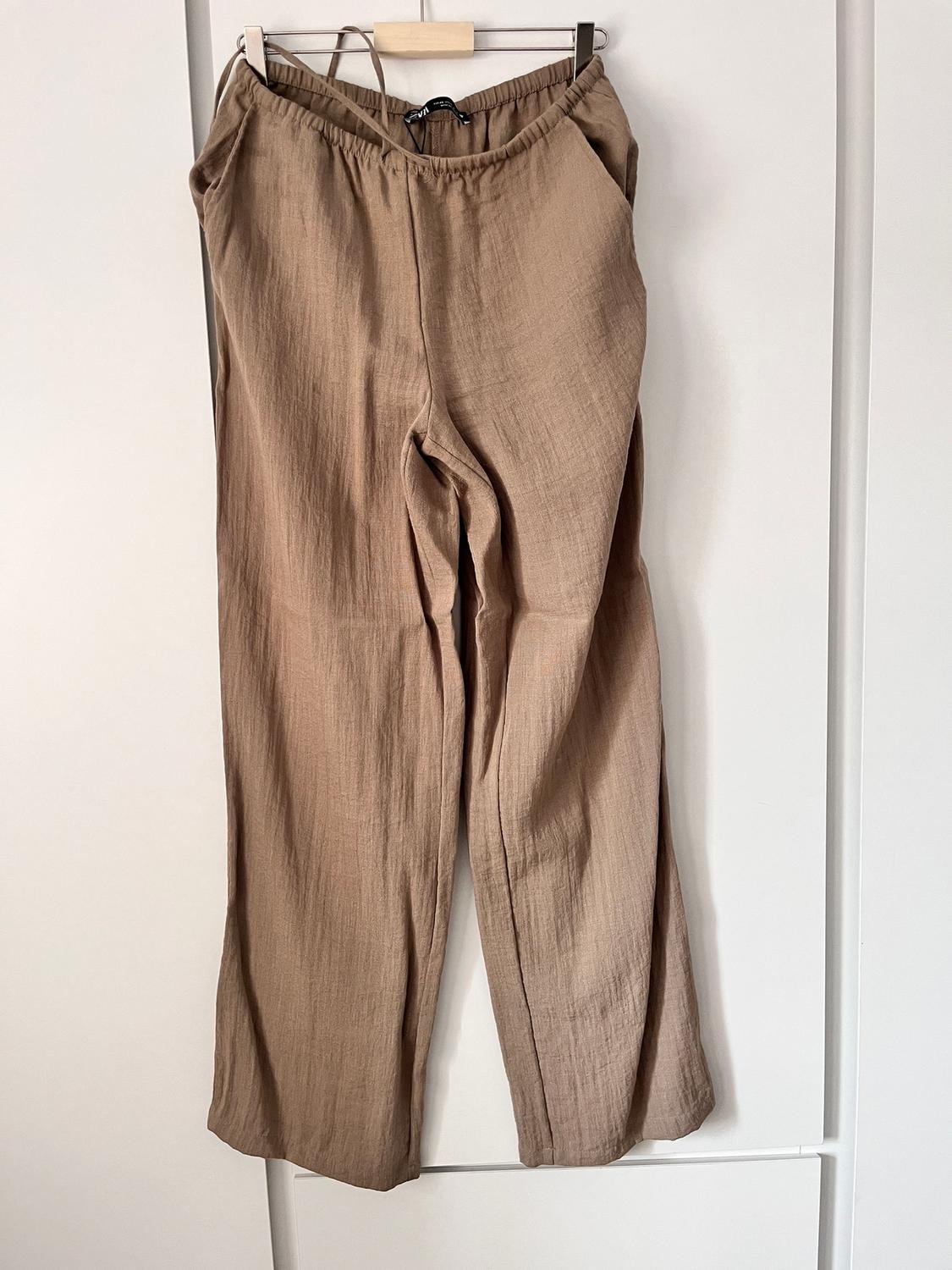 Light-coloured beige trousers hanging on a wooden hanger against a white background.
