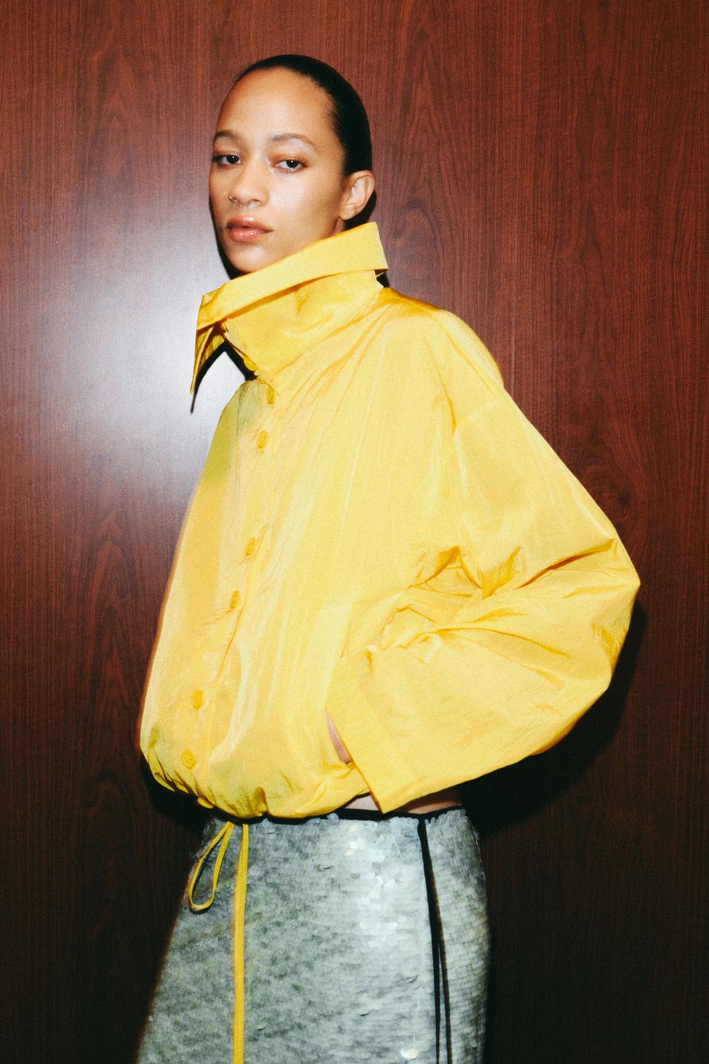 Medium shot of a yellow jacket with a high collar, long sleeves, front button fastening, side pockets, and a gathered hem.