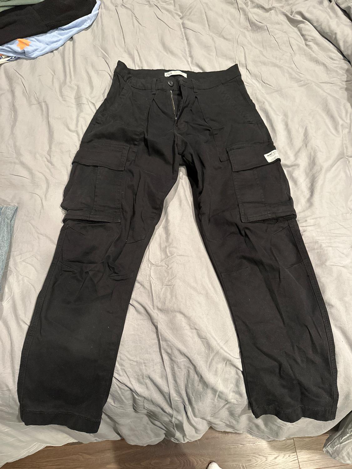 Front view of black cargo trousers with large side pockets and a visible zip.