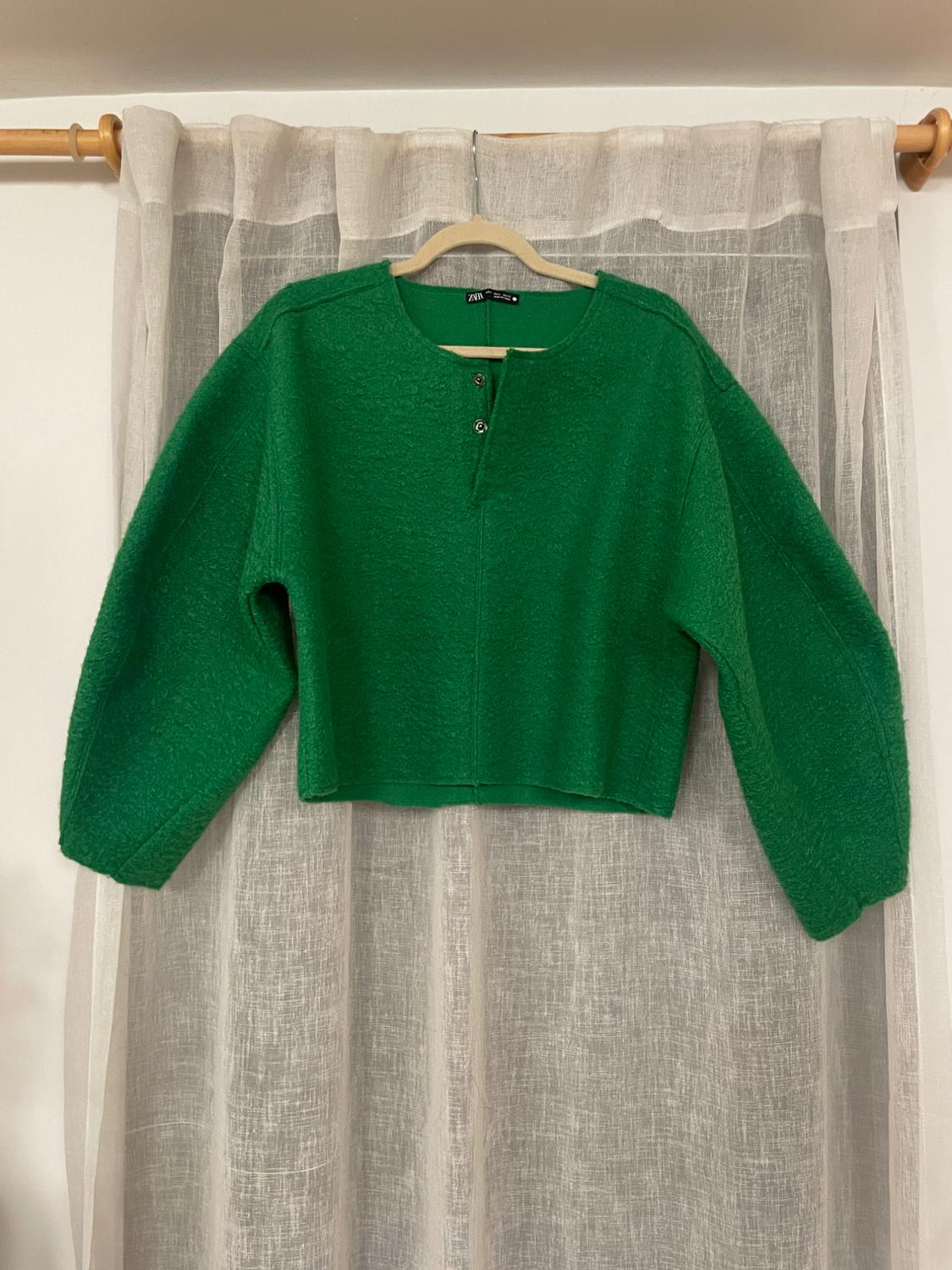 Green sweater with buttons on the collar, hanging on a wooden hanger in front of a translucent white curtain.