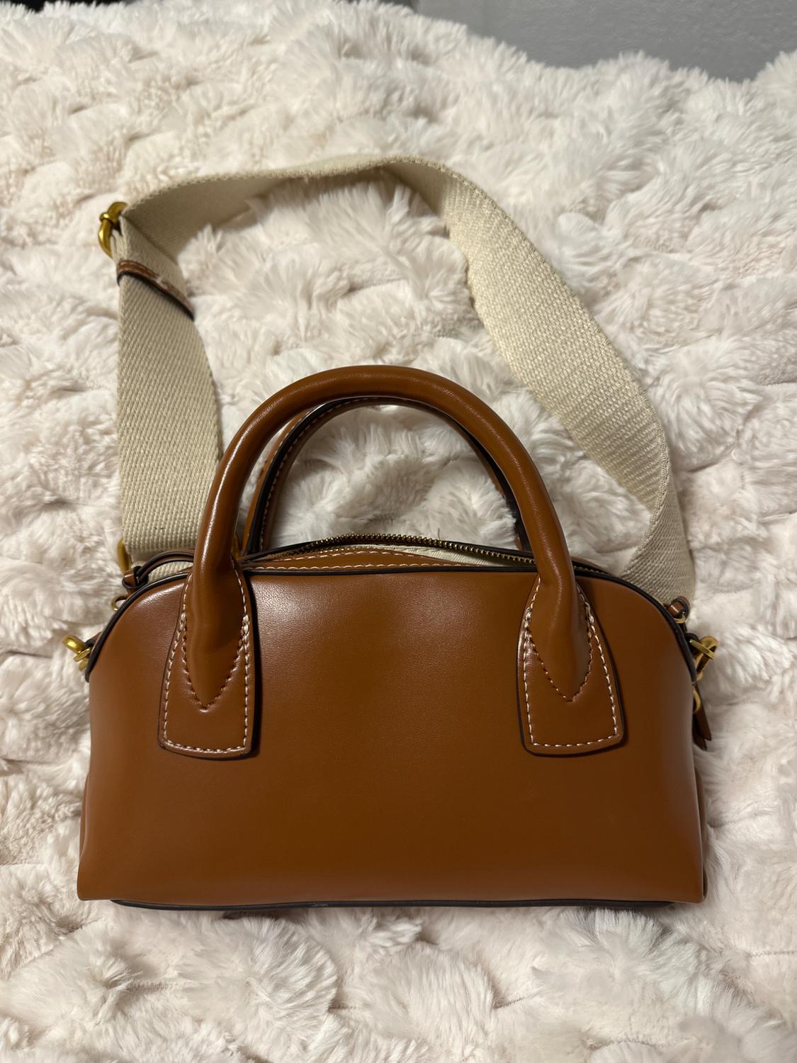Brown handbag with short handles and an adjustable beige strap on a white, fluffy fabric surface.