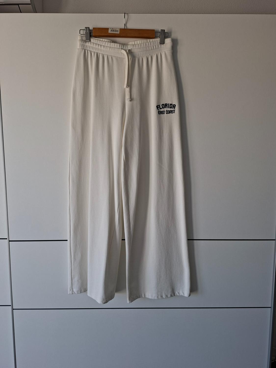 Wide white trousers hanging from a hanger in a wardrobe. They have a drawstring waist and the text “FLORIDA EAST COAST” printed in black.