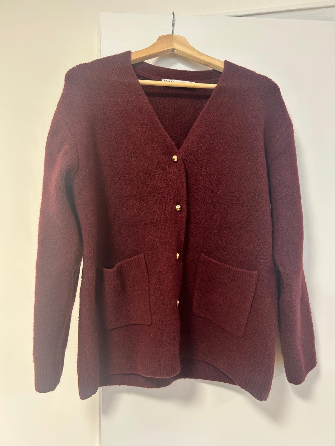Burgundy knitted cardigan with gold buttons and front pockets, hanging on a wooden hanger against a white background.