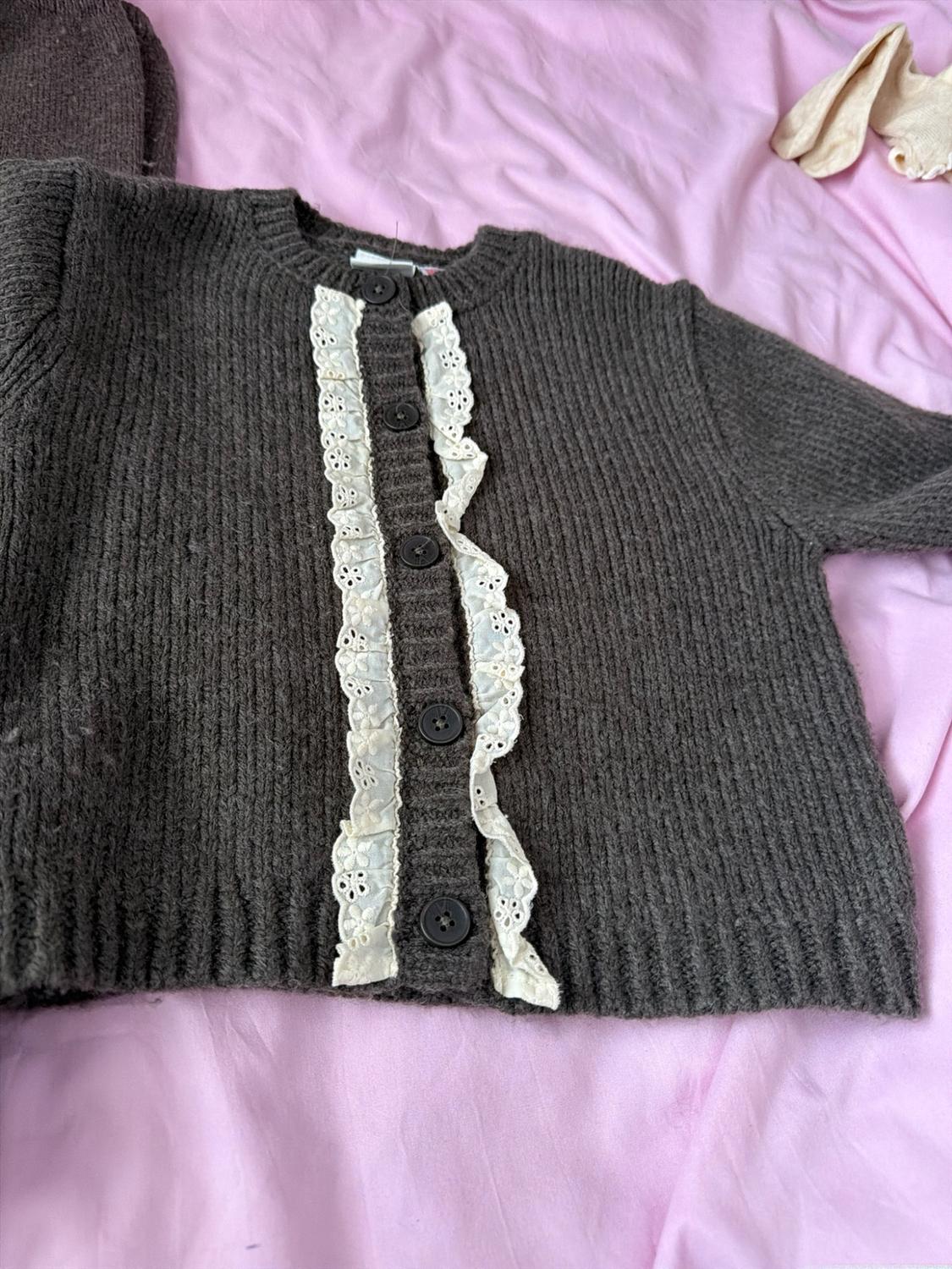 Dark brown knitted cardigan with buttons and white lace trim, laid out on a crumpled light pink fabric.