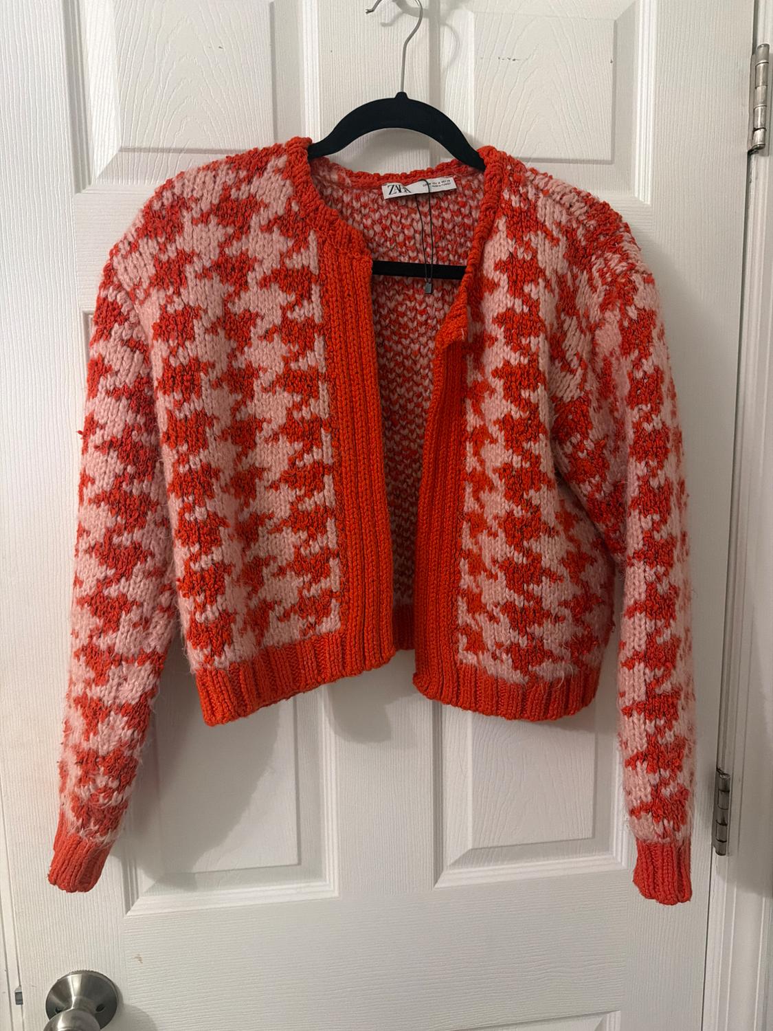 Red and white checkered knit cardigan, hanging on a black hanger over a white door.