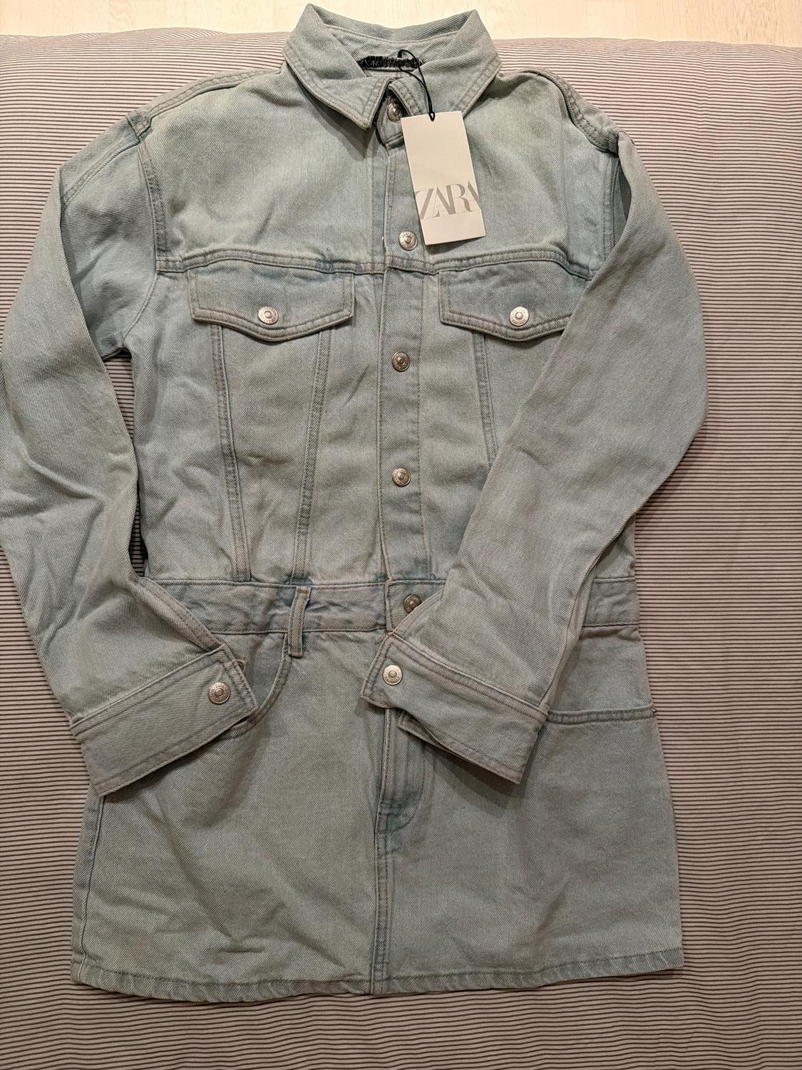 Light blue denim jacket with pockets and metal buttons, hang tag with "ZARA" text, on a striped fabric background.