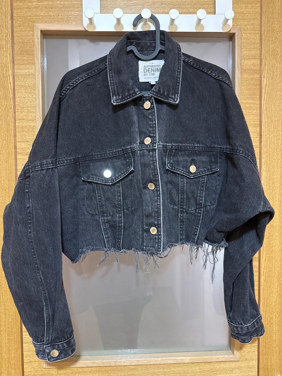 Short black denim jacket with gold buttons and frayed hem, hanging on a hanger against a light wood background.