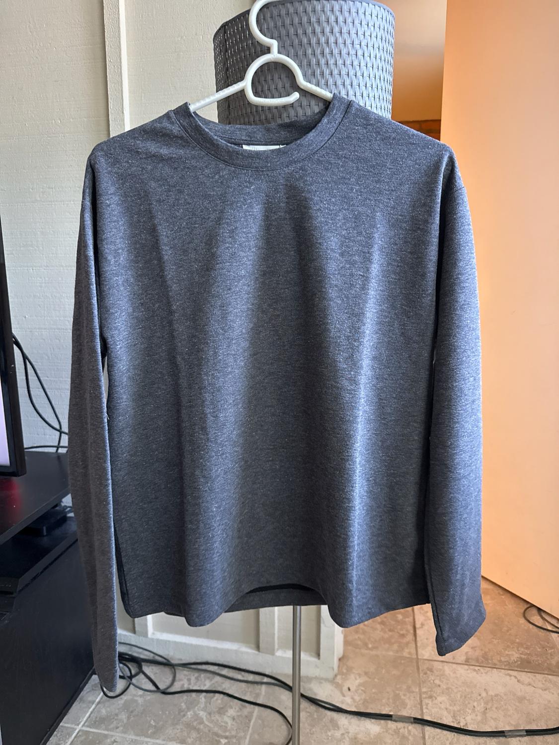 Basic long-sleeved T-shirt in dark grey, hanging on a white hanger in a casual domestic setting with a tiled floor.