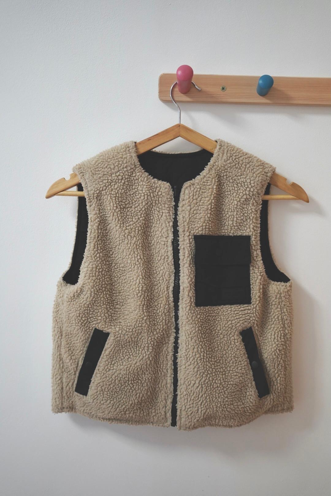Beige wool waistcoat with black pockets hanging on a wooden hanger on a wall hook with colourful knobs.