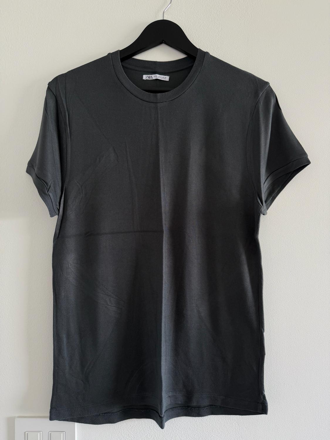 Dark gray short-sleeved t-shirt hanging on a black hanger against a white wall. The neck label reads ZARA.