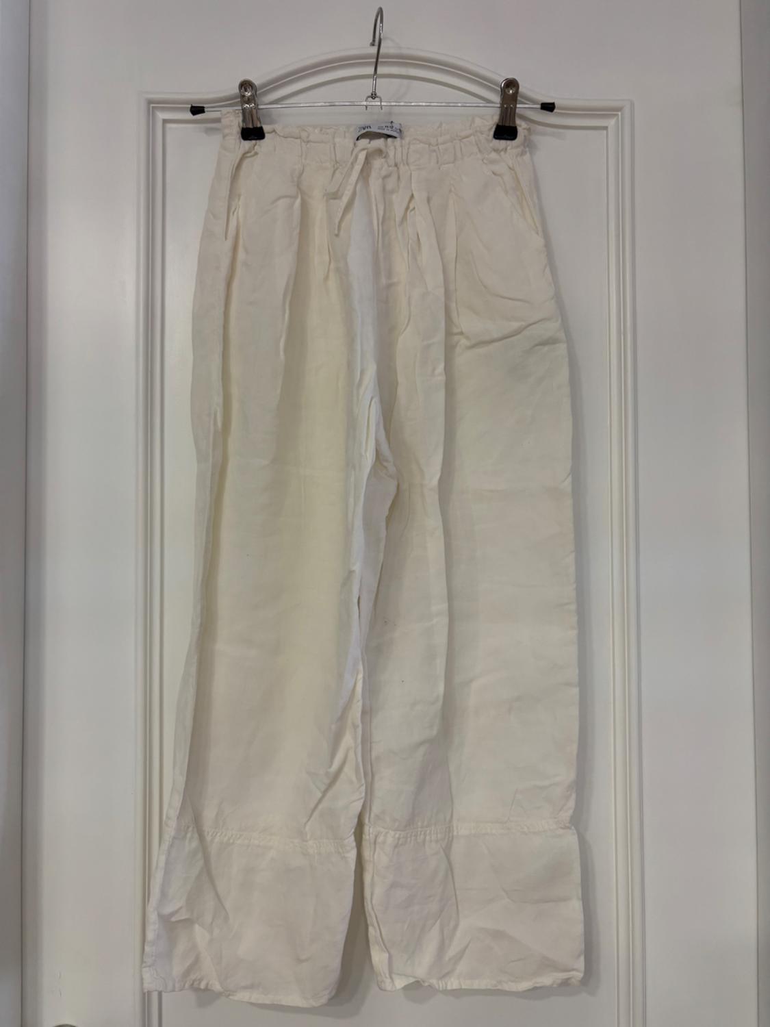 White trousers made of lightweight fabric hanging on a hanger over a white door. Loose-fitting design with adjustable elasticated waistband.