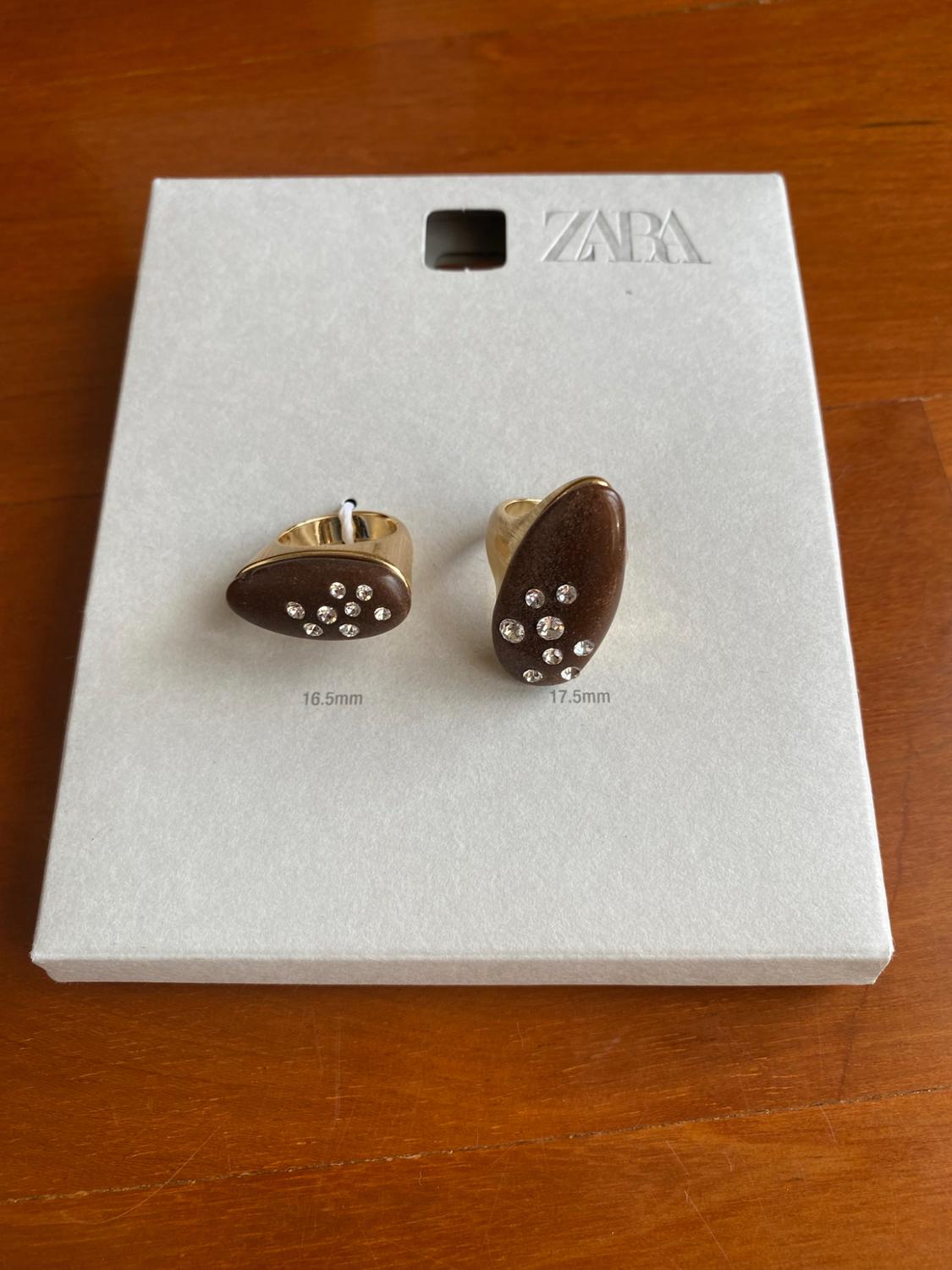 Two gold rings with a brown stone and crystals on white card. Text: ZARA, 16.5 mm, 17.5 mm. Wooden background.