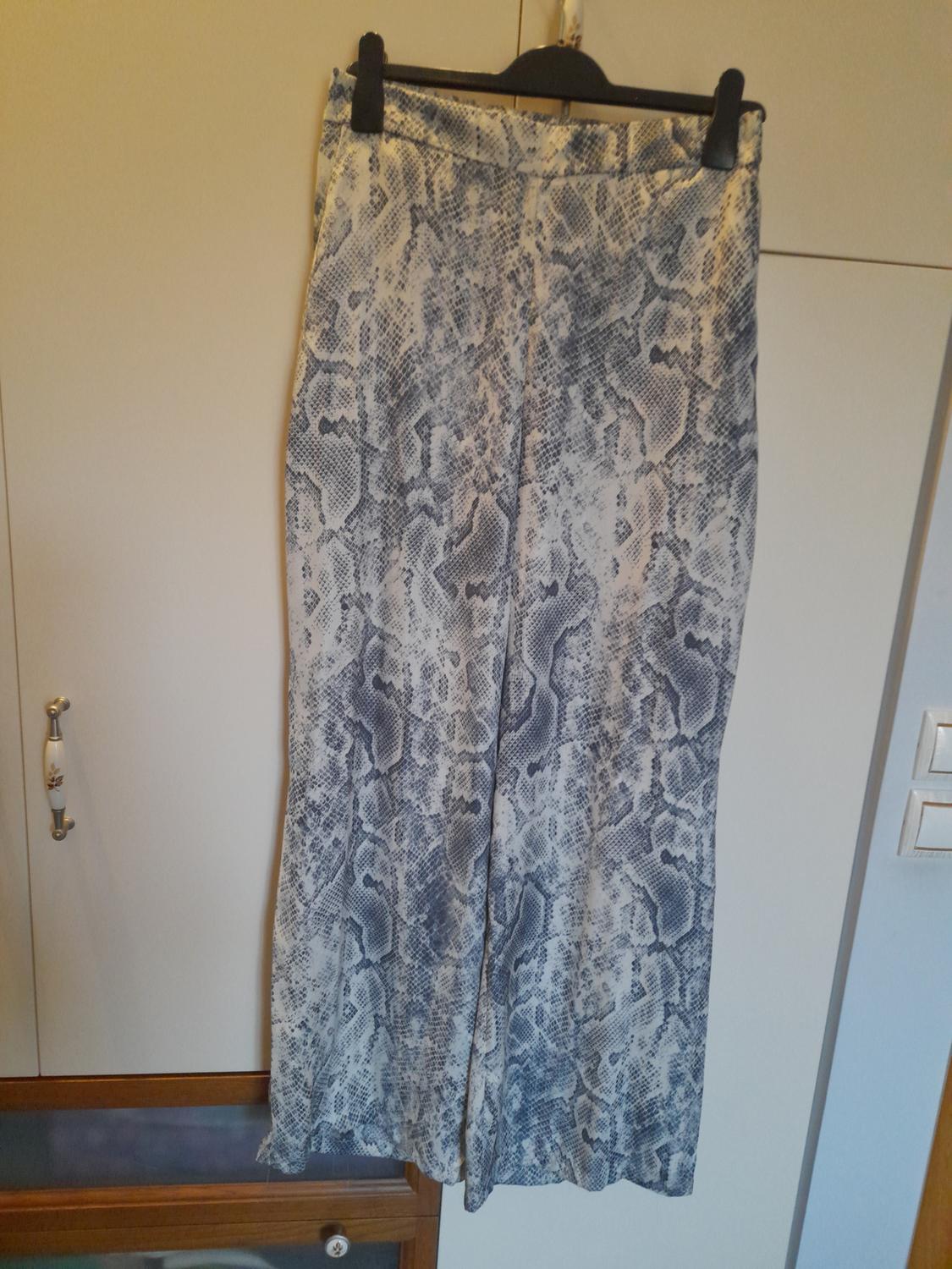 Wide-leg trousers in a grey and white snake print hanging on a black hanger in front of a white wardrobe in a relaxed setting.