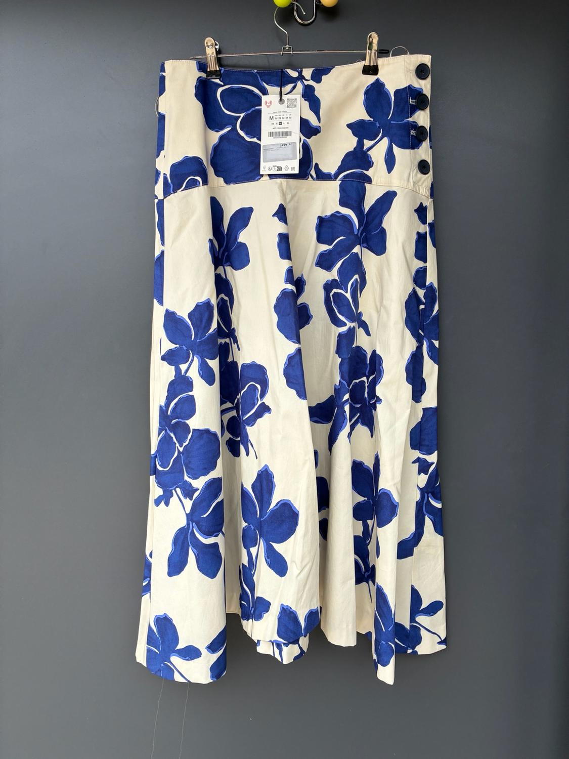 Cream-coloured long skirt with a blue floral print hanging on a hanger against a grey background. It has side buttons and a label marked M.