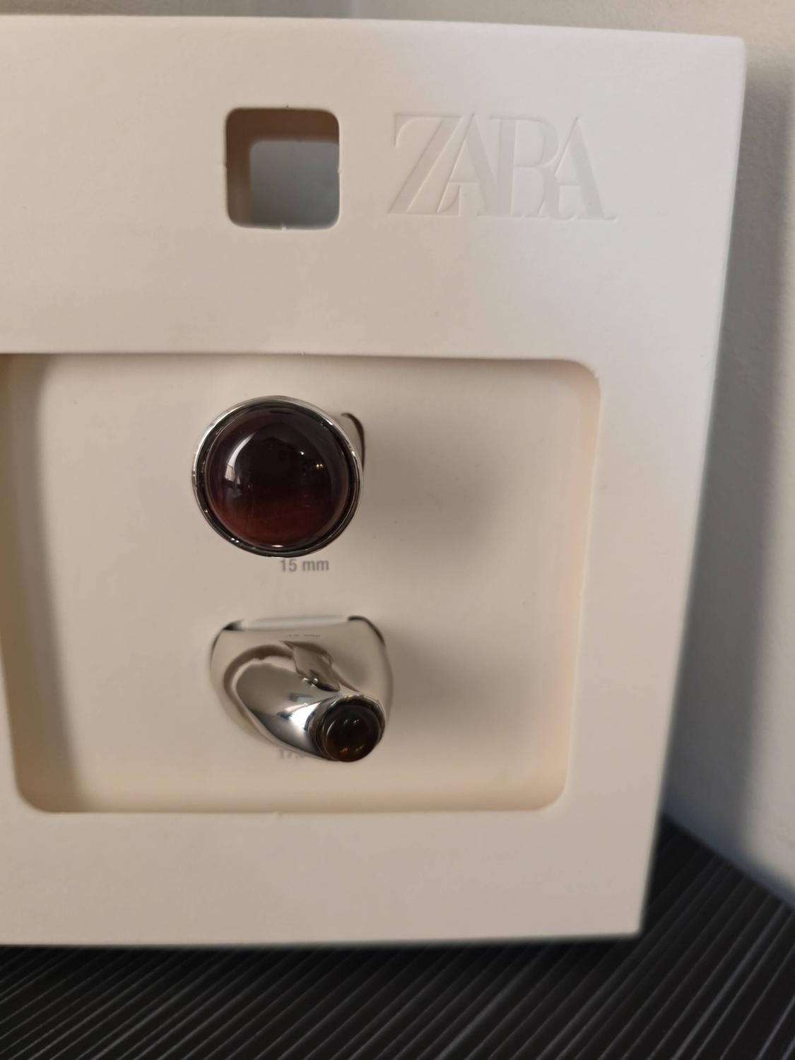 Δύο ασημένια δαχτυλίδια με σκουρόχρωμες πέτρες σε λευκή συσκευασία. Περιλαμβάνει τα κείμενα ZARA και 15 mm.