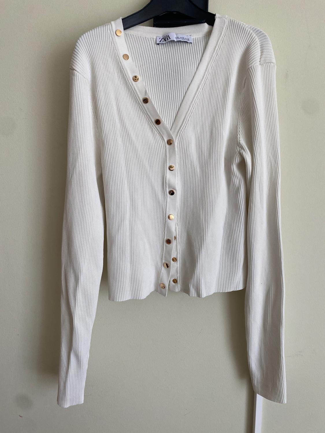 Fine-knit white cardigan with gold buttons on the front, hanging on a black hanger against a beige background.