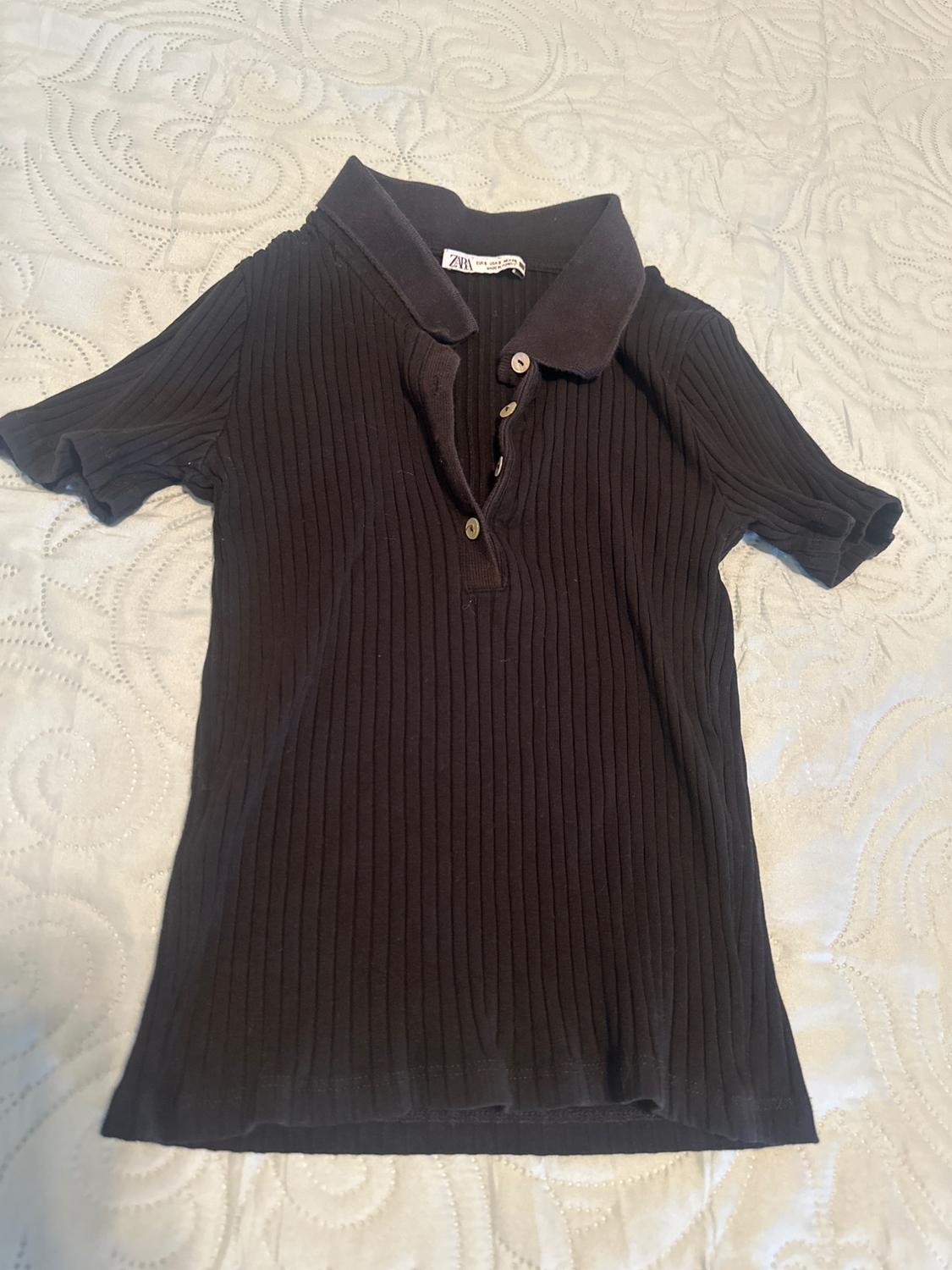 Black ribbed polo shirt with short sleeves, collar, buttons and ZARA label. Garment laid out on a light, textured bedspread.