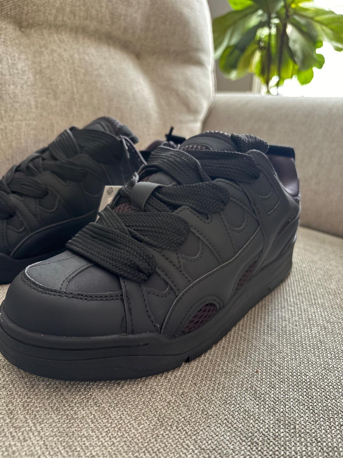 A pair of black chunky-style sneakers with thick laces and side mesh panels, on a beige sofa.