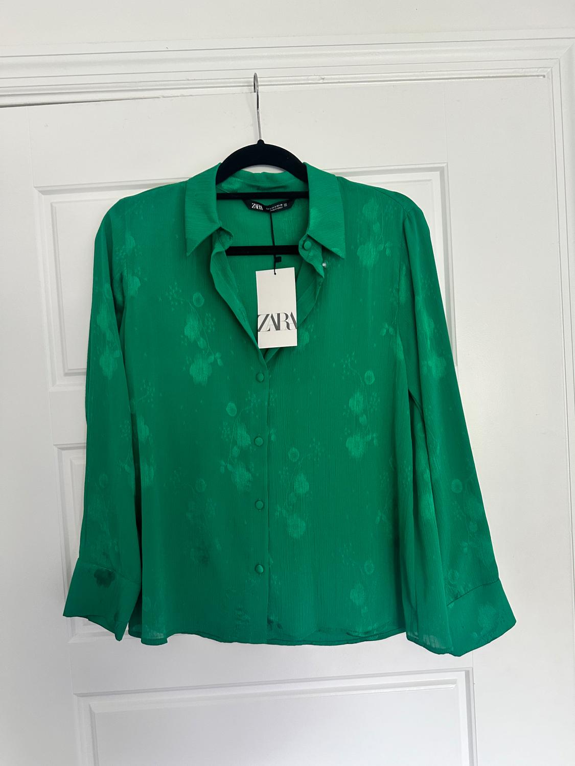 Green blouse with a satin texture and buttons down the front, hanging on a black hanger in front of a white door. Label visible with the Zara logo.
