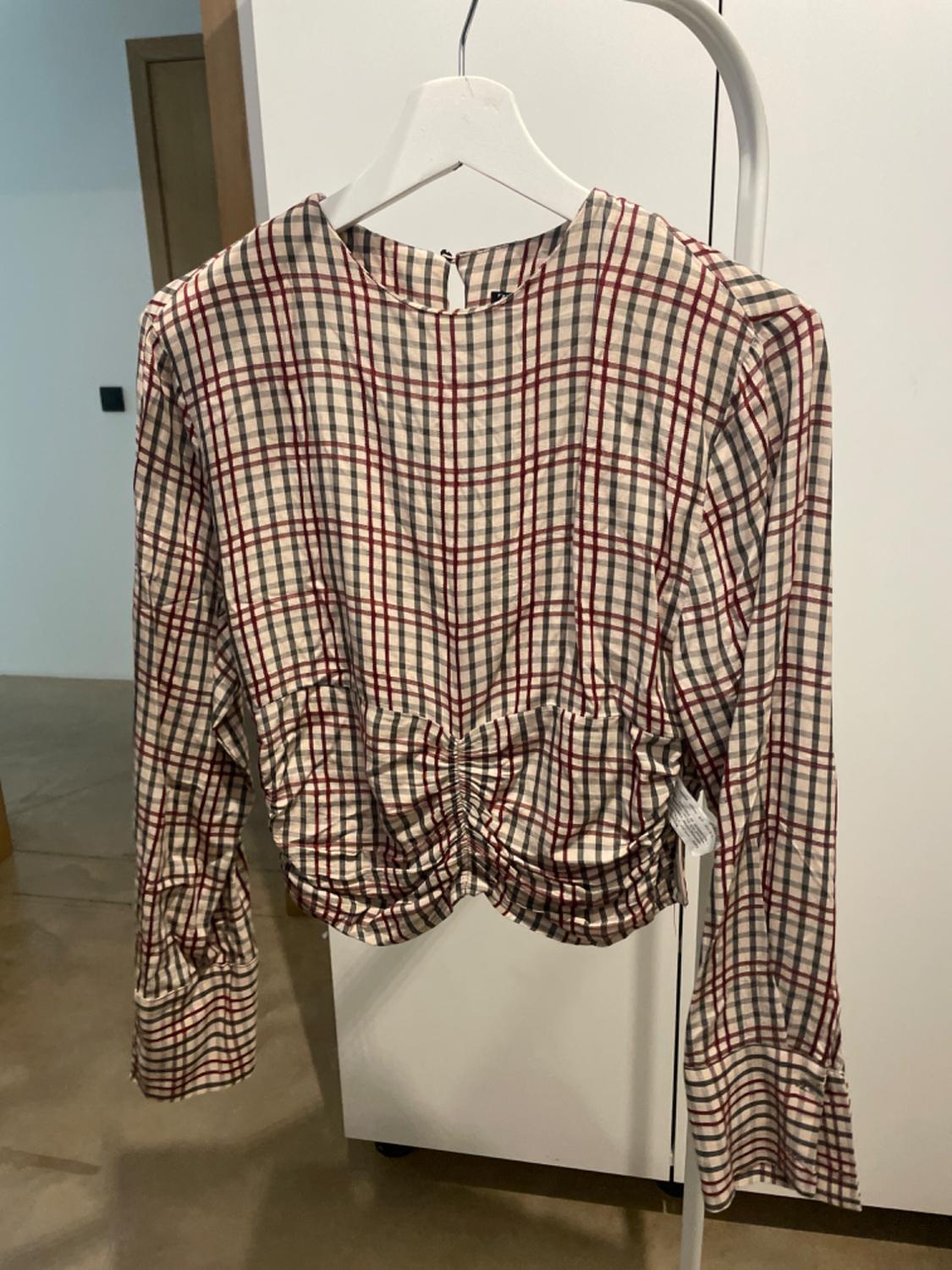 Checked blouse in beige and red tones with gathering at the waist, hanging on a white hanger against an indoor background.