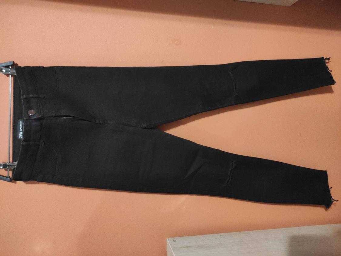 Black skinny trousers hung on a hanger against an orange wall. They have distressed details at the knees and frayed hems.