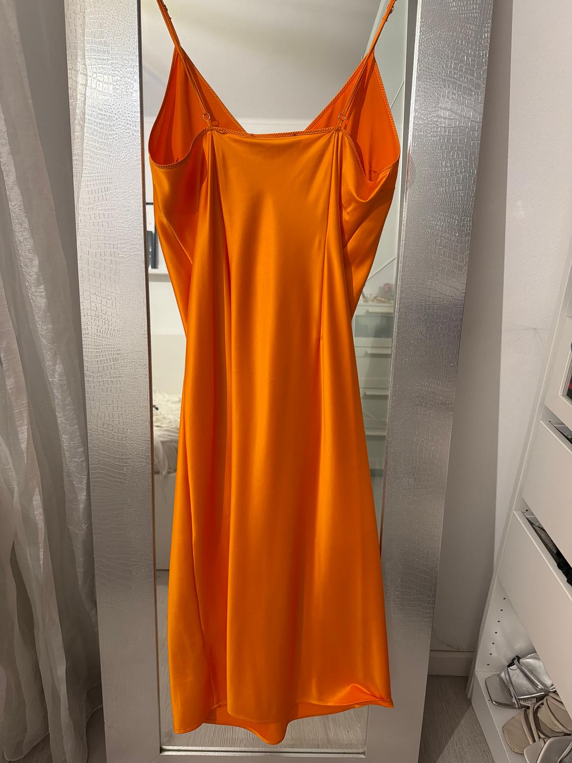 Orange spaghetti strap dress hanging on a full-length mirror in a room with white curtains and shelves.