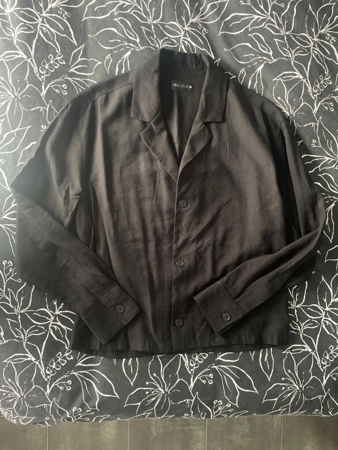 Black long-sleeved button-up shirt, on a fabric background with a white leaf design. Visible label on the collar.