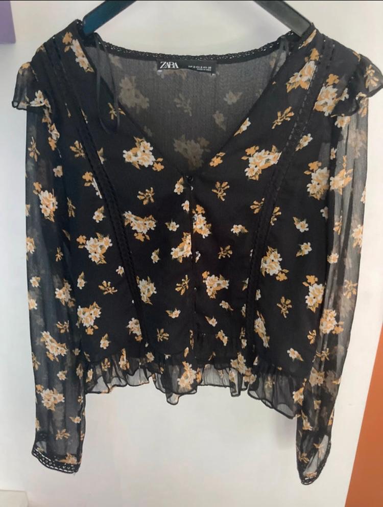 Semi-sheer black blouse with white and yellow floral print, hanging on a hanger. Visible label: "ZARA".