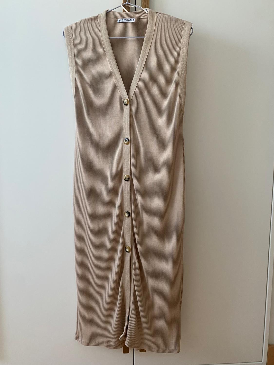 Sleeveless beige dress with gold buttons, hanging on a hanger against a white background.