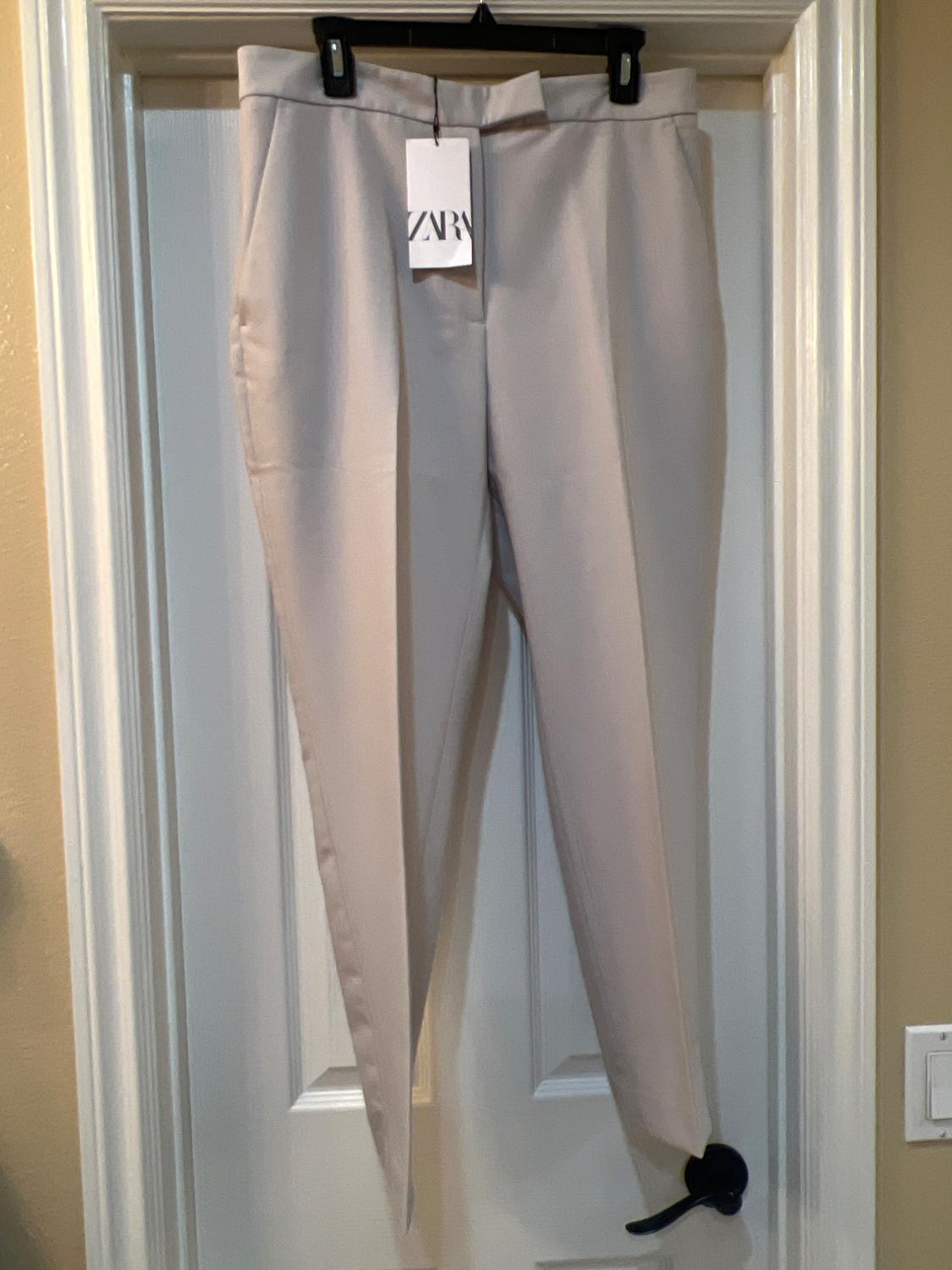 Beige trousers hanging on a white door with a label that says "ZARA". The door has a black handle.