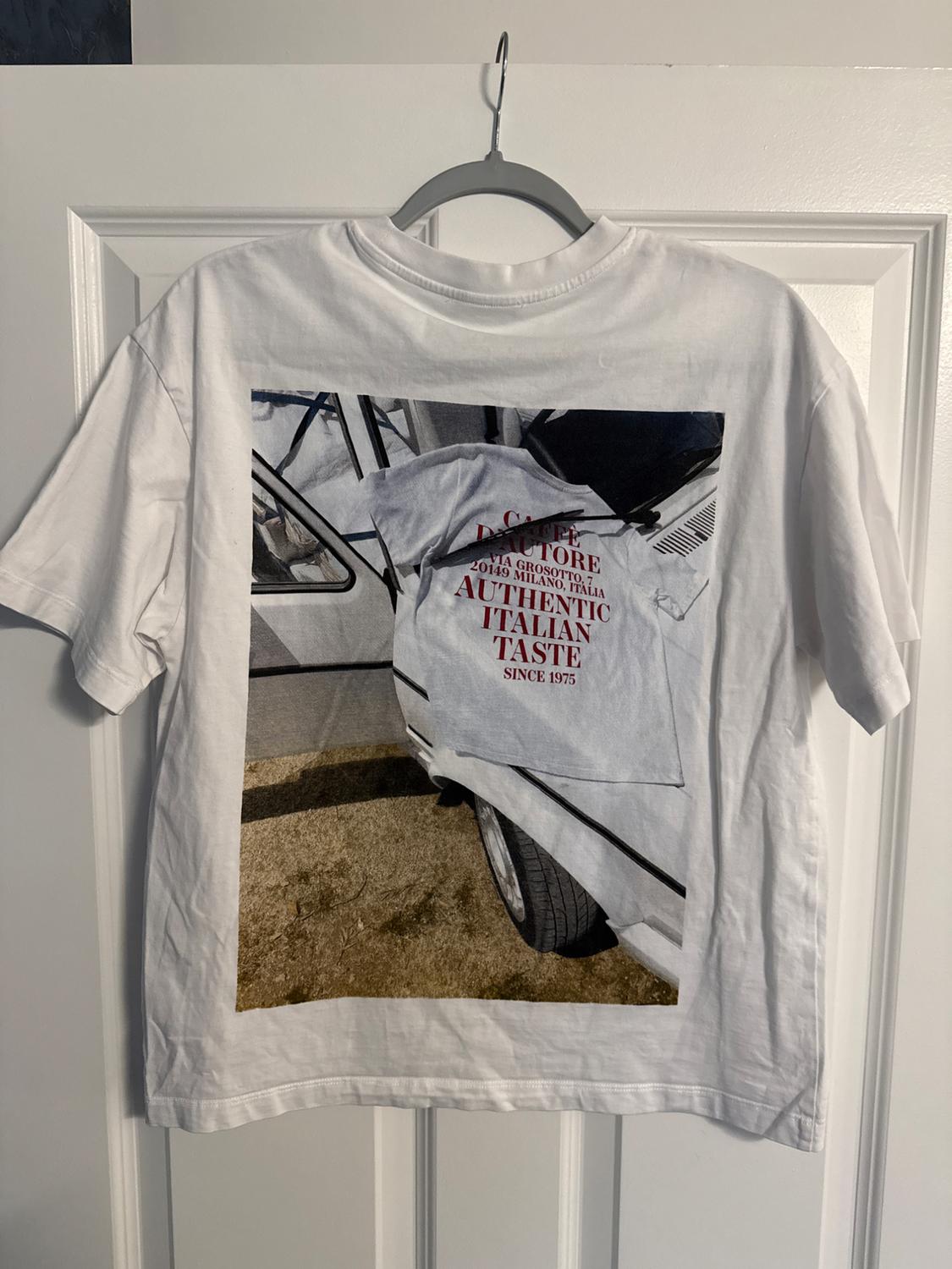White t-shirt hanging with image of covered car and text: "CAFFÈ A. TORE 20145 MILANO, ITALIA AUTHENTIC ITALIAN TASTE SINCE 1975".