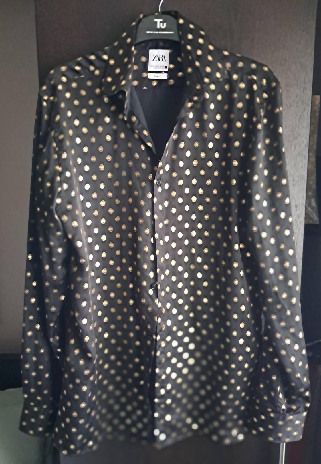 Black shirt with gold polka dots hanging on a black hanger with the "ZARA" label visible. Dark background.