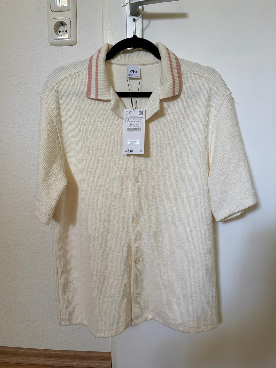 Cream-coloured, short-sleeved, textured shirt with a pink striped collar, hanging on a white door. Text: ZARA, S.