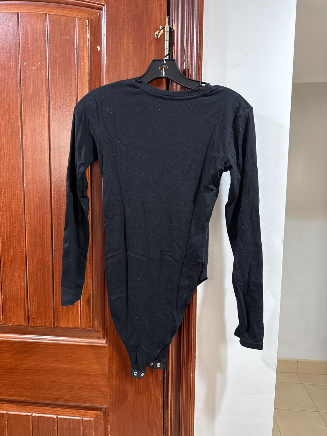 Long-sleeved black bodysuit hanging on a hanger over a wooden door. The garment has a snap closure at the bottom.