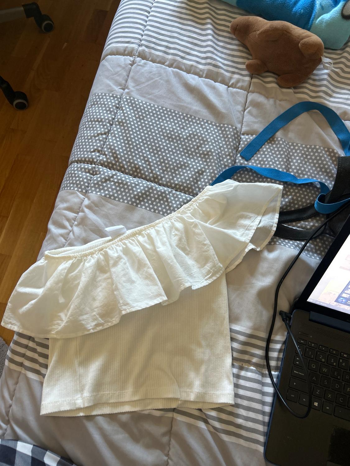 White one-shoulder frill blouse on a bed with a striped and polka dot quilt, next to a laptop and a brown soft toy.
