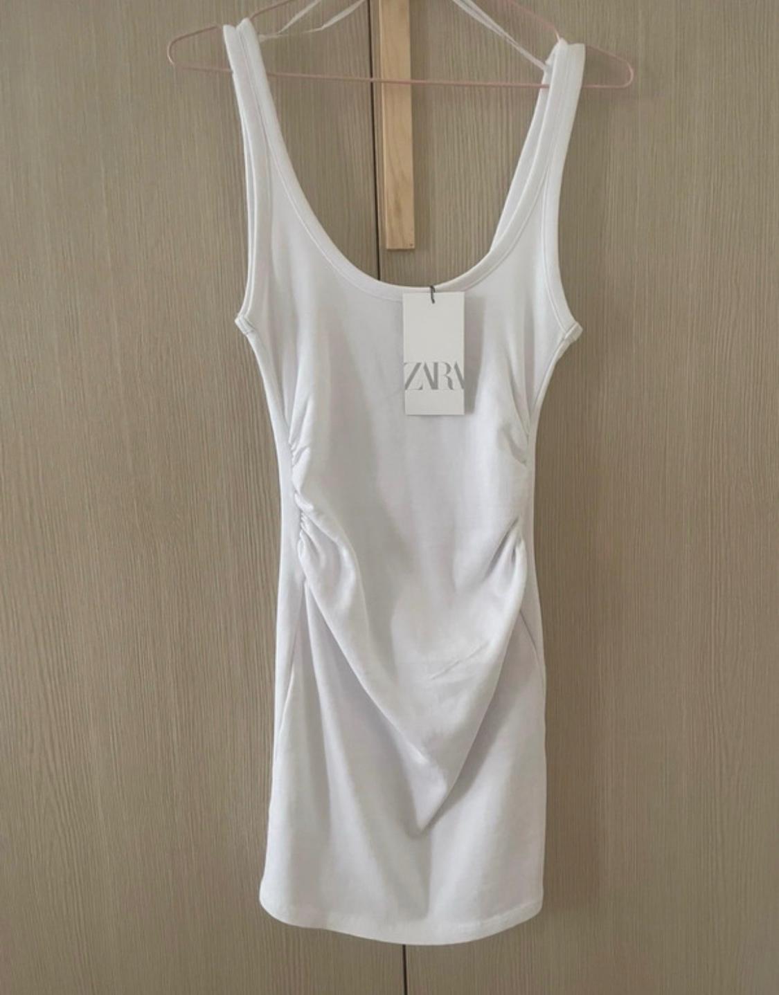 Sleeveless white dress with gathered detail, hanging on a hanger against a wooden background. Label with ZARA text.