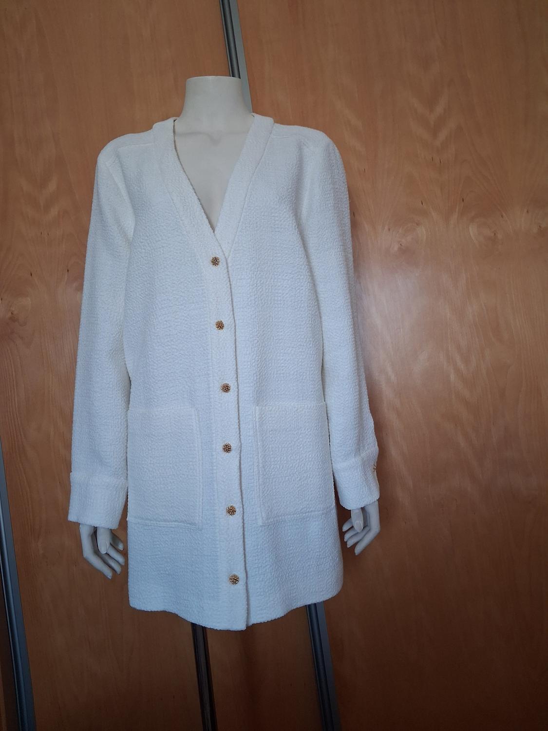 Mannequin in a long white jacket with gold buttons and large pockets, against a light wooden background.