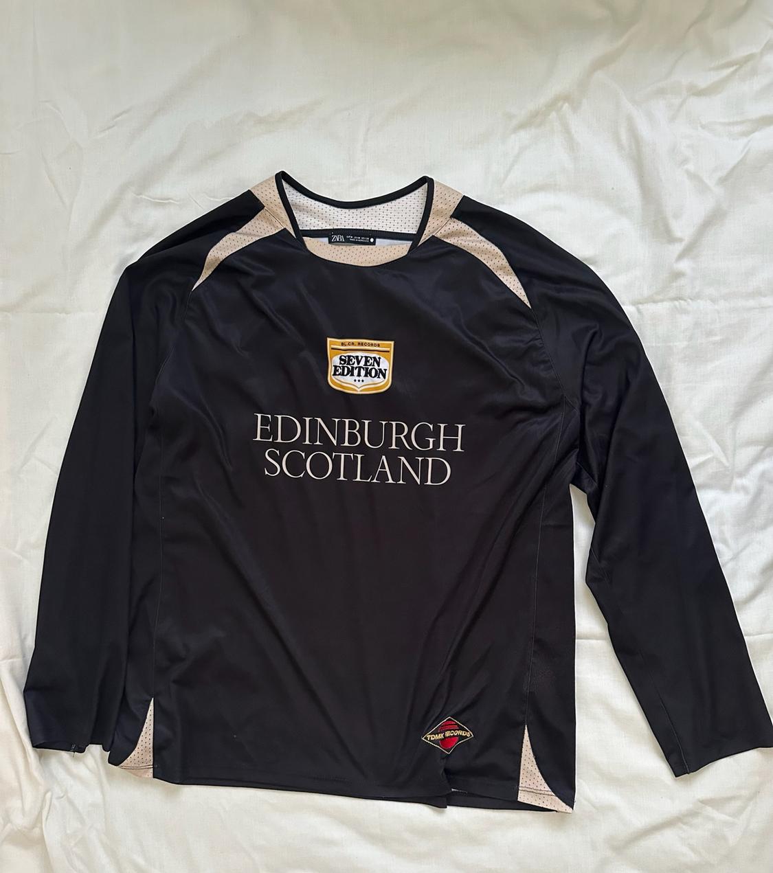 Black long-sleeved T-shirt on white fabric. Beige details and "SEVEN EDITION EDINBURGH SCOTLAND" text. Sporty and relaxed style.