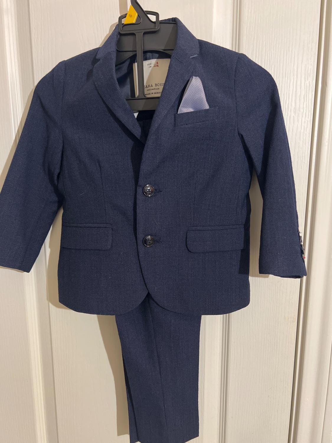 Blue children's suit hanging on a black hanger, with a white handkerchief in the chest pocket, against a white door background.