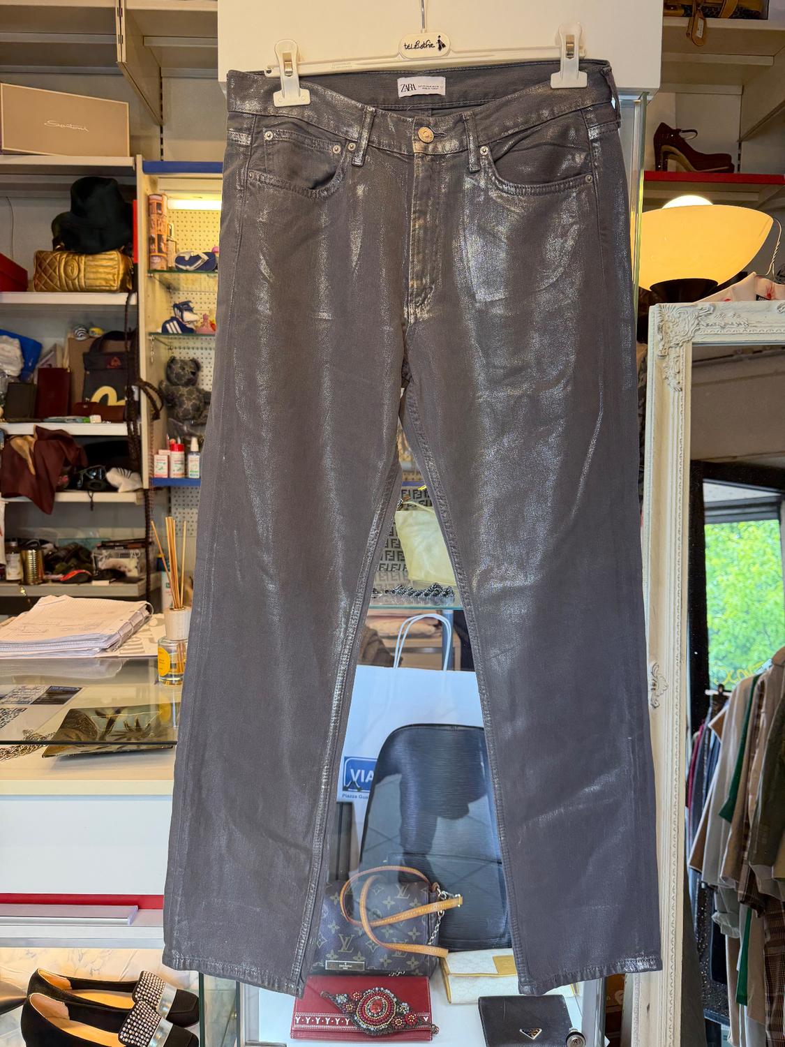 Shiny grey trousers hanging on a hanger, with a shop background displaying various handbags, shoes, and accessories.