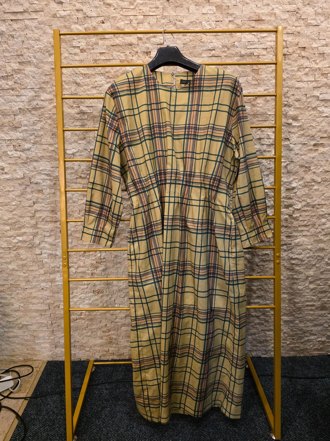 Long plaid dress in brown and green tones hanging on a gold clothes rack, against a beige stone wall background.