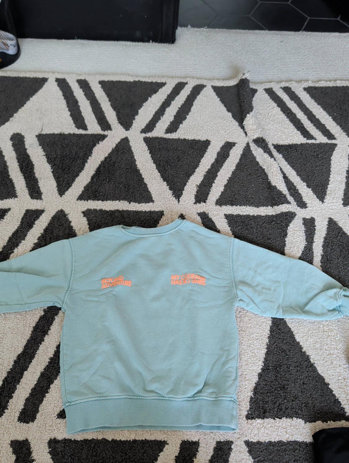 Light green sweatshirt on a geometric rug with the orange text “BEYOND ADVENTURE” and “MY COURAGE HAS NO LIMITS”.